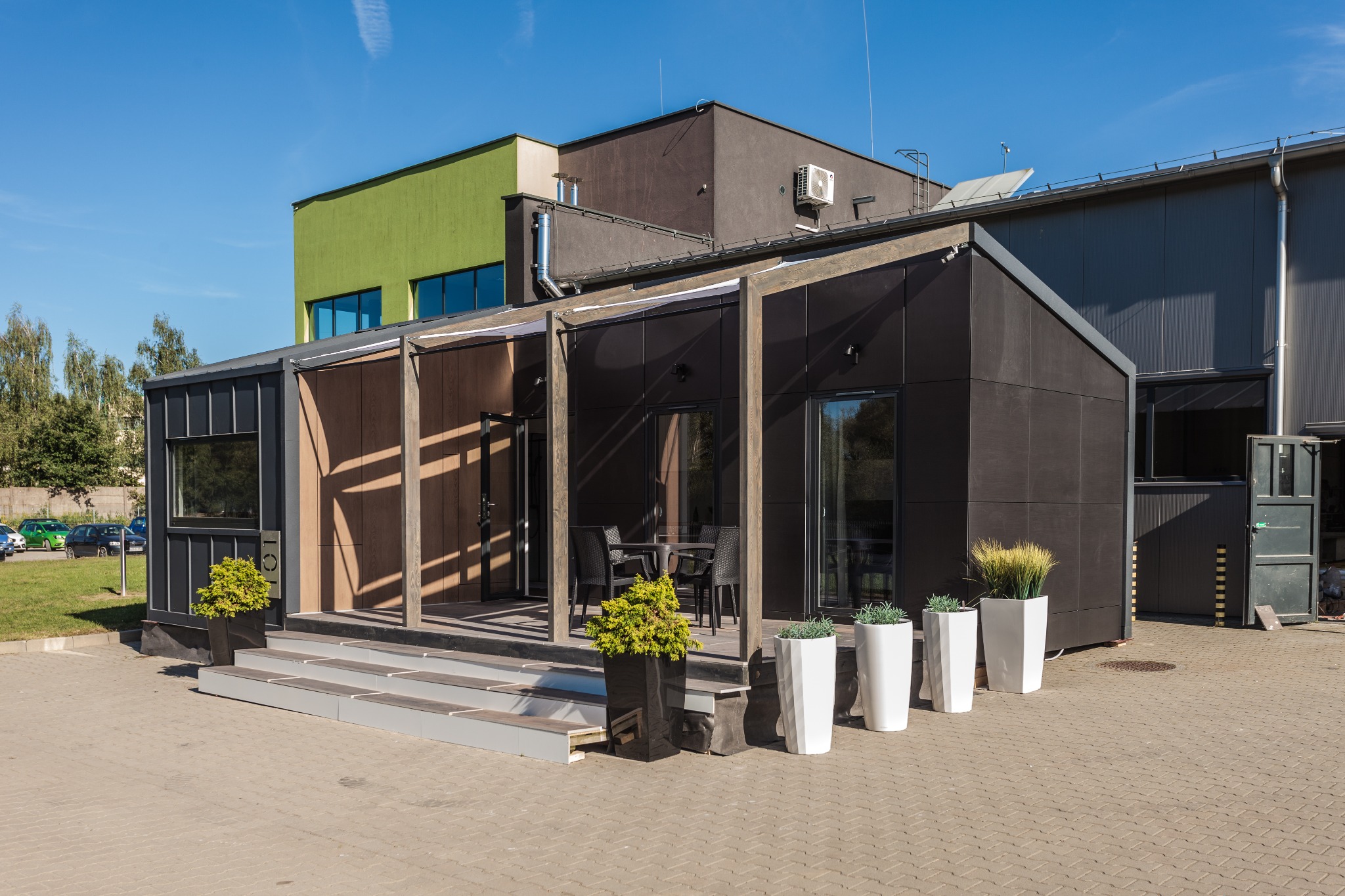 Nowoczesny dom modułowy z ciemną elewacją, tarasem z meblami ogrodowymi i ozdobnymi donicami, na tle budynków przemysłowych w słoneczny dzień.