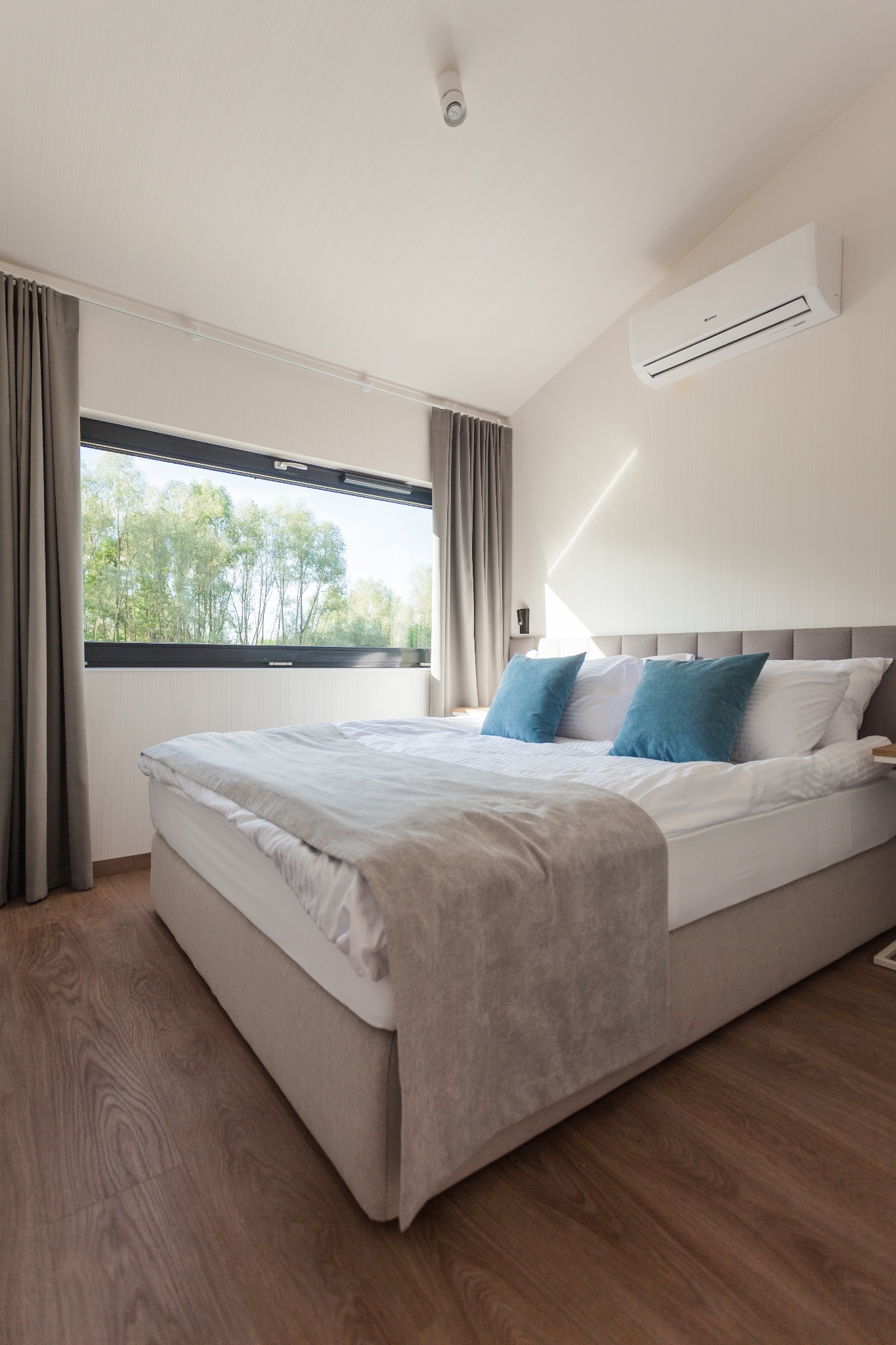 Jasna sypialnia z dużym oknem z widokiem na drzewa, łóżko z szarym zagłówkiem i turkusowymi poduszkami, klimatyzator na ścianie, drewniana podłoga.
