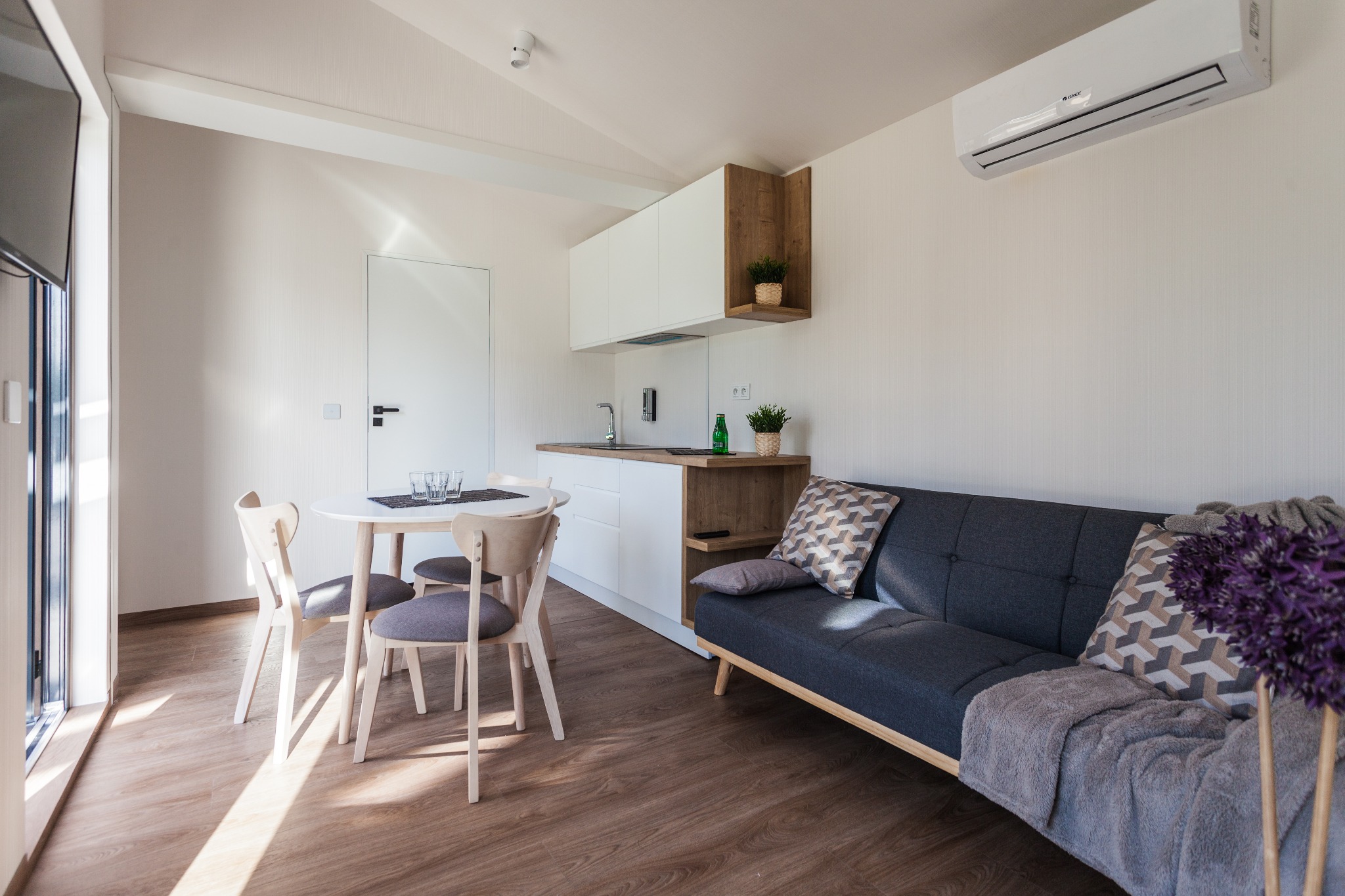 Jasne wnętrze pokoju dziennego z aneksem kuchennym: szary narożnik z poduszkami, okrągły stół z krzesłami, biała zabudowa kuchenna z drewnianymi akcentami, klimatyzator na ścianie.