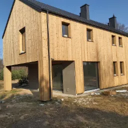 Nowoczesny dom z elewacją z jasnego drewna w trakcie budowy, widoczne niedokończone detale i materiały budowlane na terenie posesji.