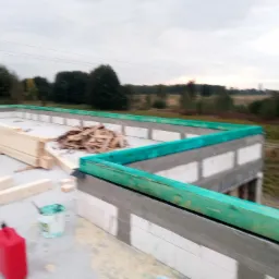 Wznoszone ściany domu z betonu komórkowego w Międzyborowie, zabezpieczone zieloną, drewnianą belką. Na fundamencie widoczne drewniane elementy konstrukcyjne i wiadro z farbą.