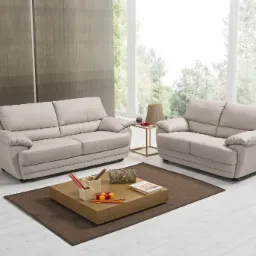 Dwa beżowe, skórzane komplety wypoczynkowe w jasnym salonie z dużym oknem i drewnianym stolikiem kawowym.