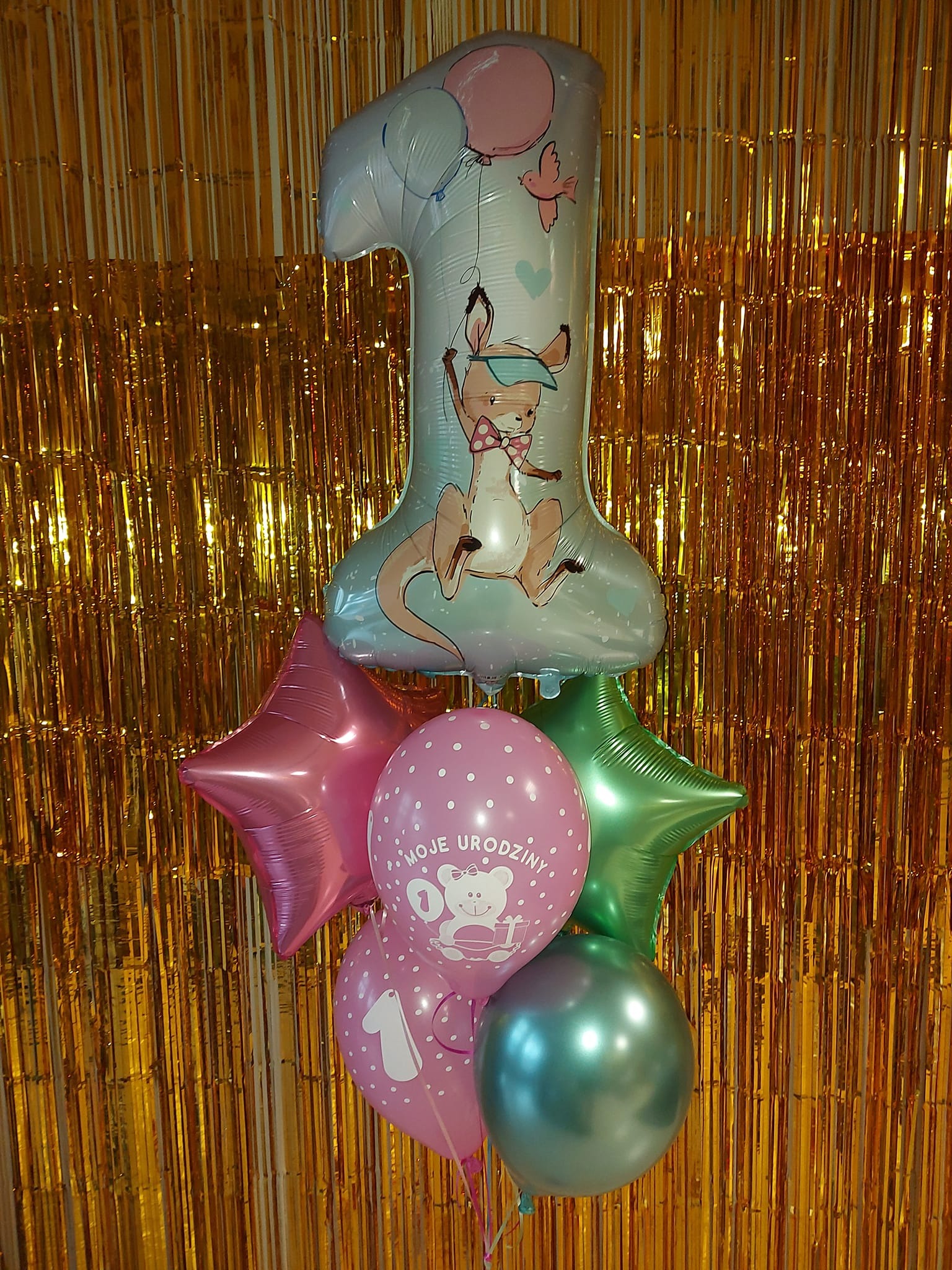 Dekoracja balonowa na pierwsze urodziny z cyfrą '1', kangurkiem, gwiazdkami i napisem 'Moje Urodziny' na tle złotego, błyszczącego kurtynowego tła.