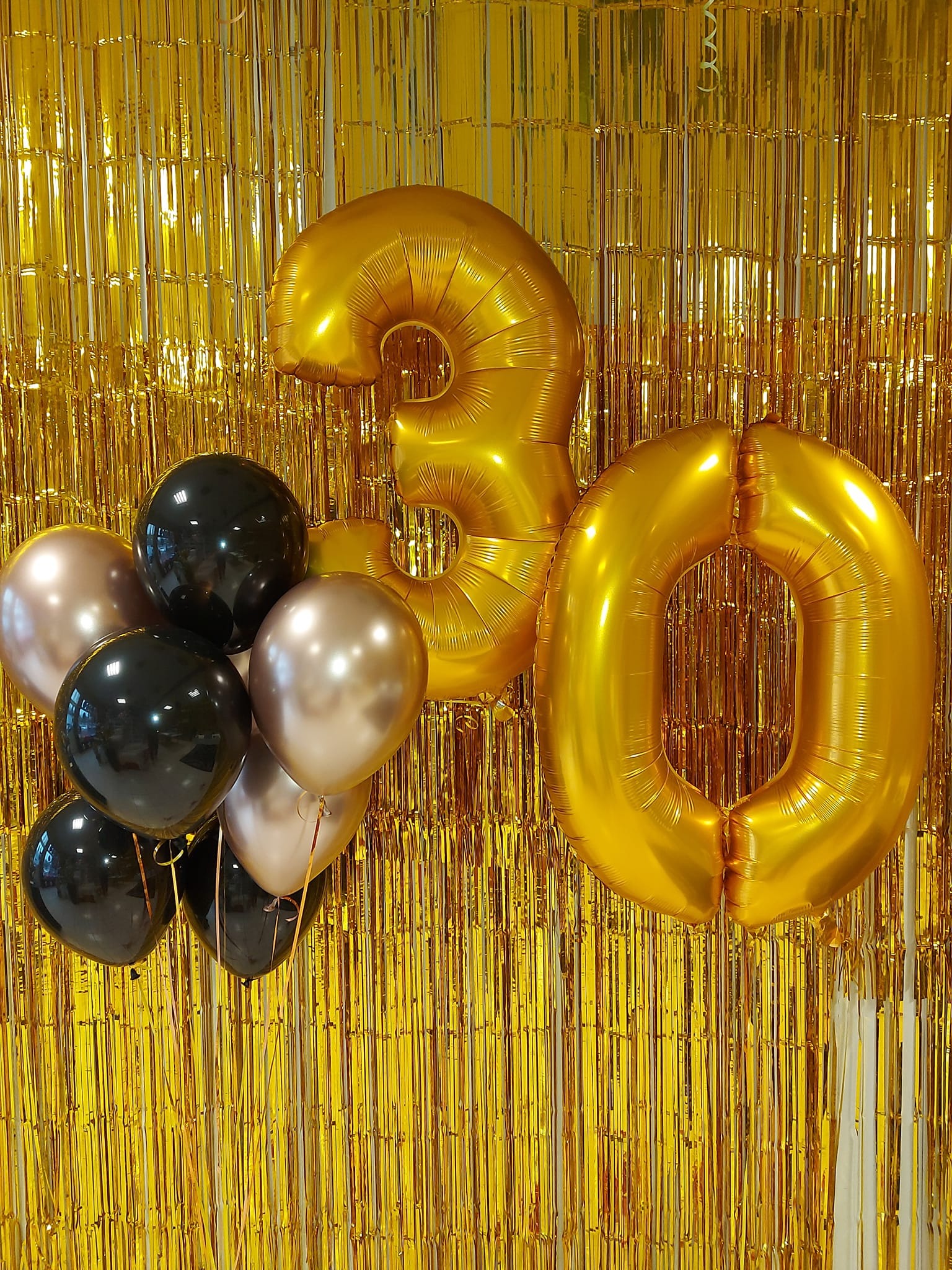 Złote balony foliowe w kształcie cyfr 30 oraz bukiet czarnych i złotych balonów na tle błyszczącej, złotej kurtyny tworzą dekorację urodzinową.