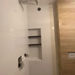 Świeżo wykafelkowana łazienka z wnęką z półkami w ścianie, armatura zabezpieczona folią, widoczne płytki imitujące drewno na jednej ze ścian.