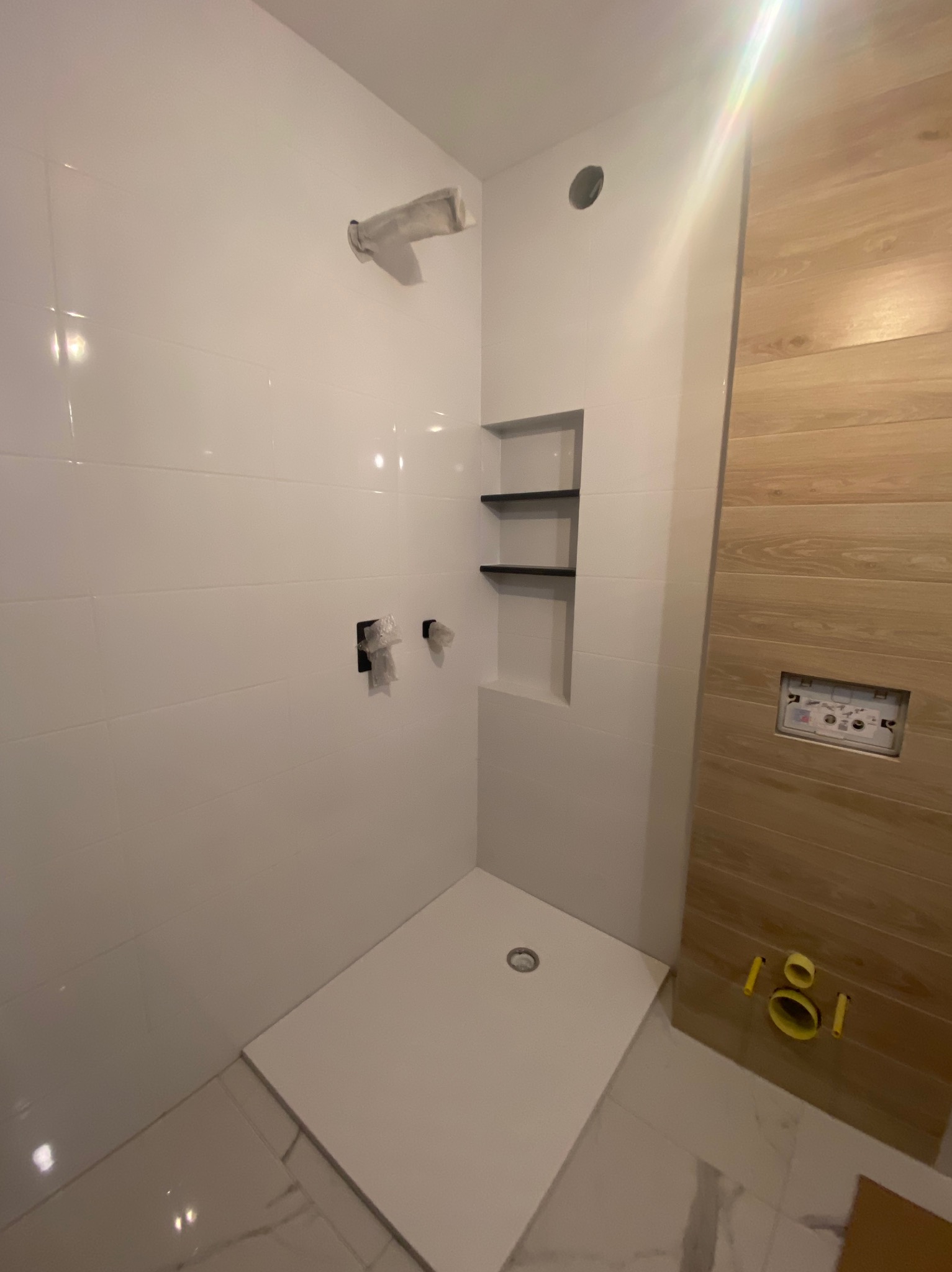 Niedokończona kabina prysznicowa z białymi płytkami, drewnopodobną ścianą z ukrytą spłuczką i wnęką z półkami, widoczne zabezpieczenia na baterii.
