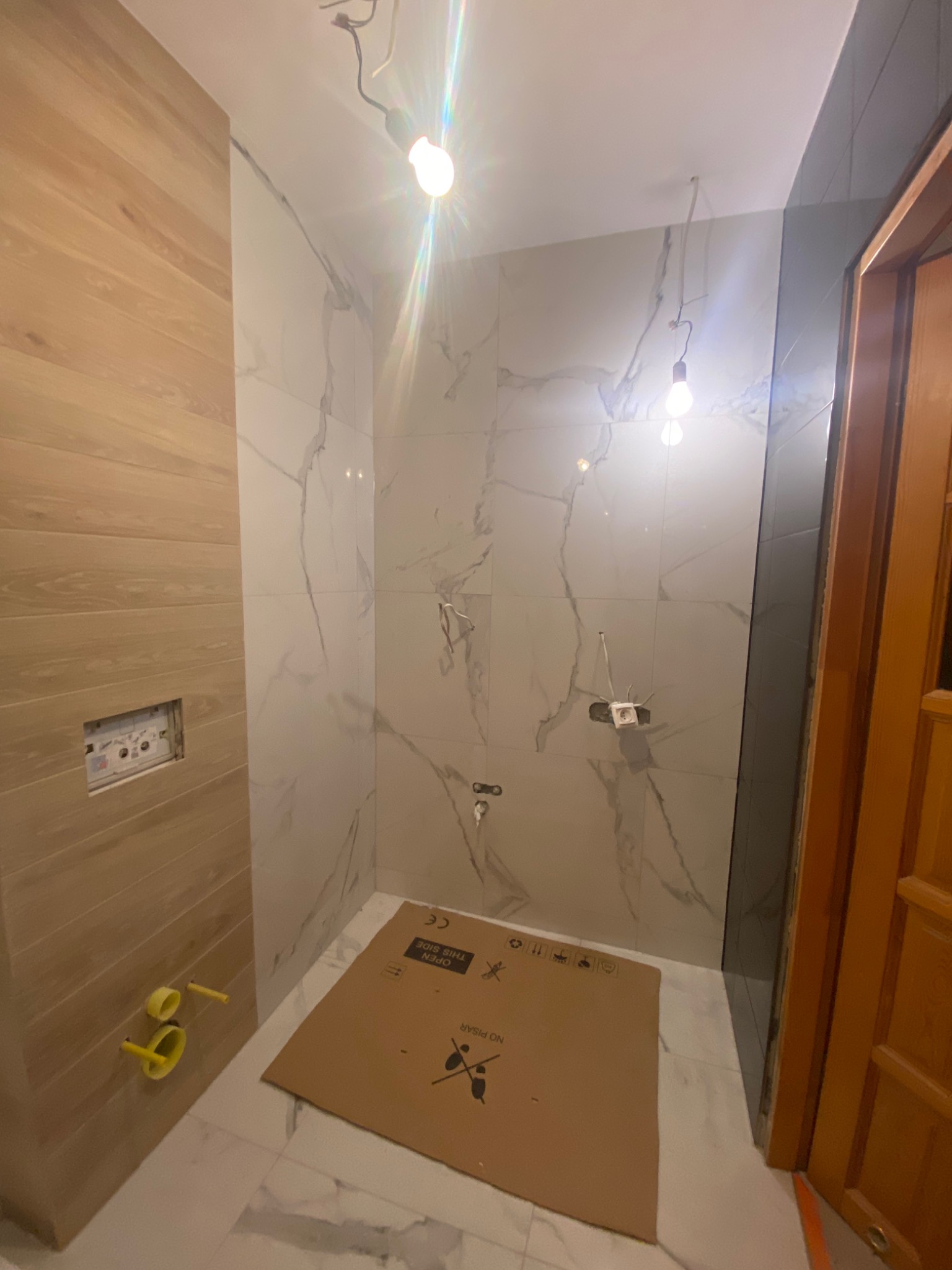 Wnętrze łazienki w trakcie remontu, z widocznymi płytkami imitującymi marmur na ścianach, drewnopodobną okładziną, niezabezpieczonymi przewodami elektrycznymi, żółtymi rurami instalacji wodnej...