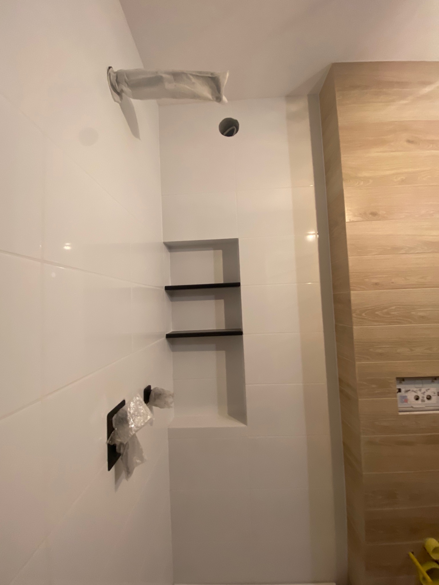 Świeżo wykafelkowana łazienka z wnęką z półkami w ścianie, armatura zabezpieczona folią, widoczne płytki imitujące drewno na jednej ze ścian.