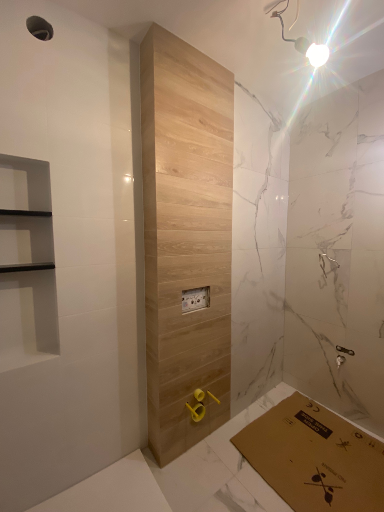 Wnętrze łazienki w trakcie prac wykończeniowych; widoczna drewnopodobna okładzina pionowa, białe płytki imitujące marmur, przygotowane instalacje, kartonowe zabezpieczenie podłogi.