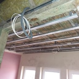 Seb- Bud - Montaż metalowego stelaża sufitu podwieszanego w pomieszczeniu z widocznymi instalacjami elektrycznymi i fragmentami ścian w trakcie prac wykończeniowych.