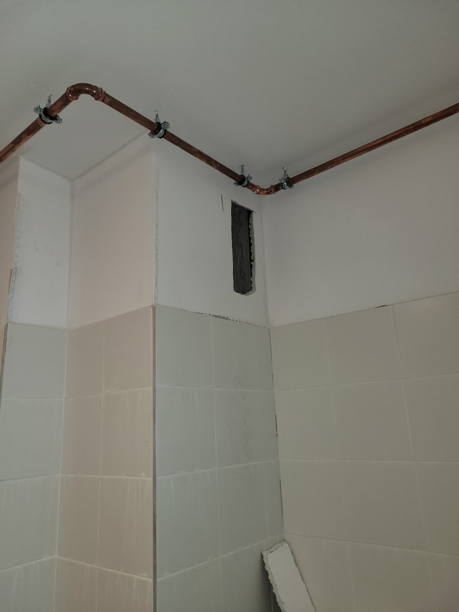 Miedziane rury instalacji CO zamocowane do sufitu i ściany w pomieszczeniu z częściowo położonymi białymi płytkami. Widoczny otwór w ścianie.