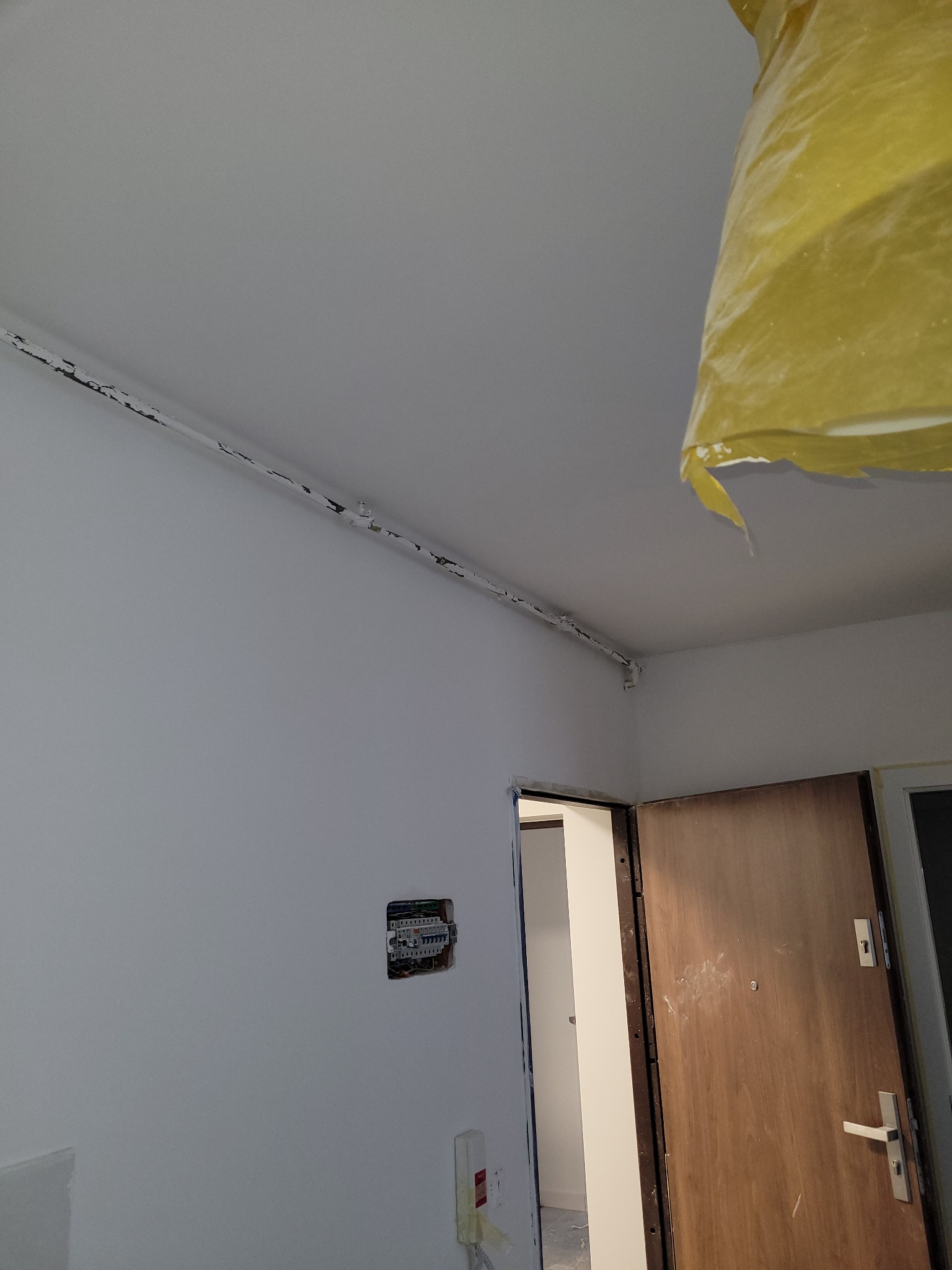 Wnętrze pokoju w trakcie odświeżania: świeżo pomalowane ściany na biało, widoczna instalacja elektryczna w ścianie, zabezpieczona folią część sufitu oraz fragment drzwi wejściowych.