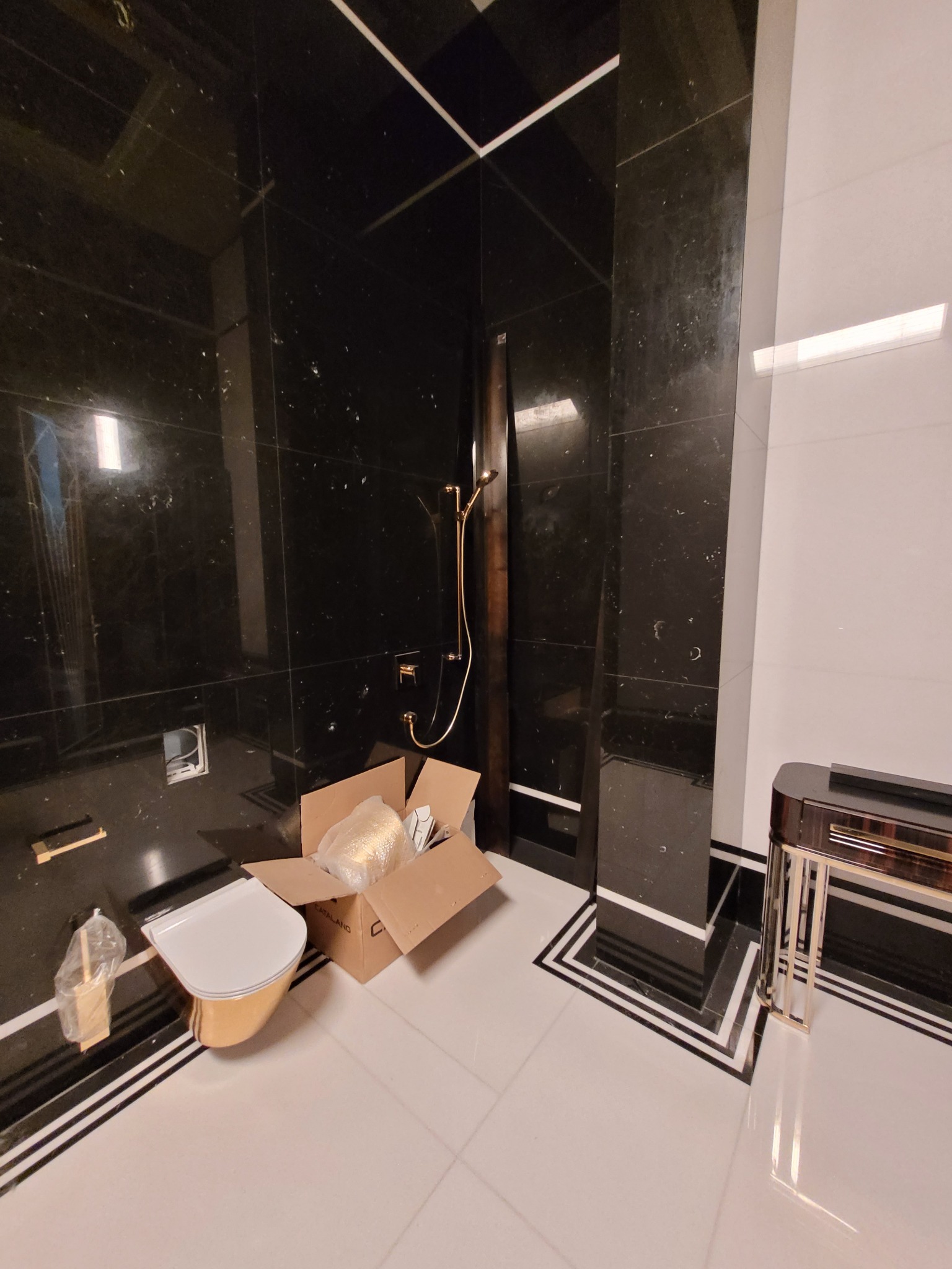 Nowa, złota toaleta Catalano w kartonie, umieszczona w łazience z czarnymi, marmurowymi płytkami i białymi płytkami podłogowymi z czarnymi obramowaniami, obok złotej słuchawki prysznicowej.