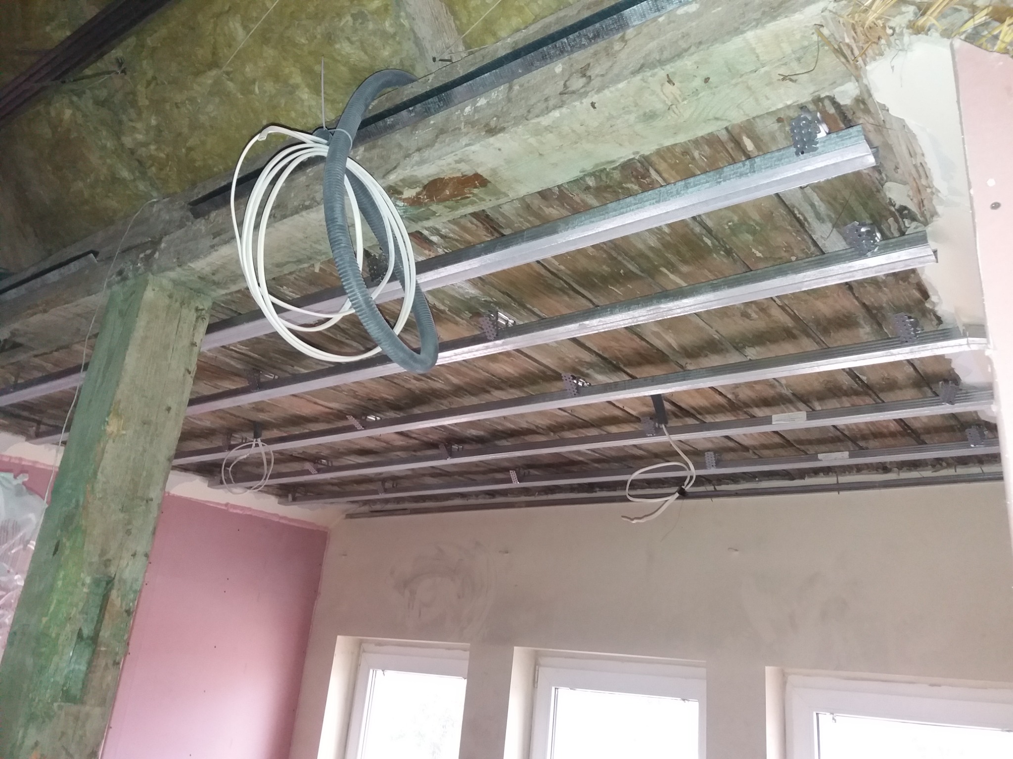 Montaż metalowego stelaża sufitu podwieszanego w pomieszczeniu z widocznymi instalacjami elektrycznymi i fragmentami ścian w trakcie prac wykończeniowych.