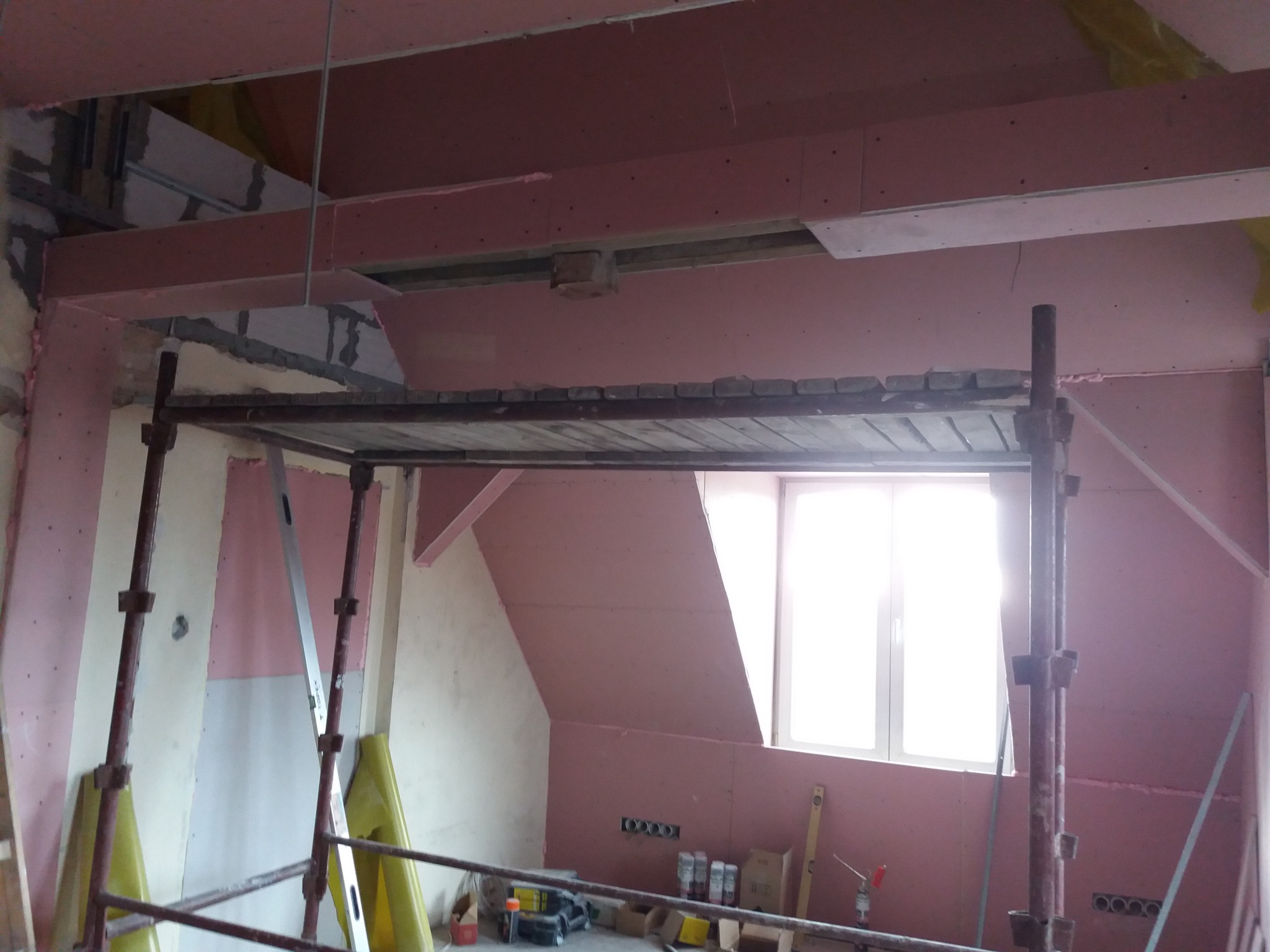 Wnętrze poddasza w trakcie prac wykończeniowych: ściany i sufit pokryte płytami gipsowo-kartonowymi w kolorze różowym, widoczne rusztowanie, narzędzia i materiały budowlane, okno w dachu.