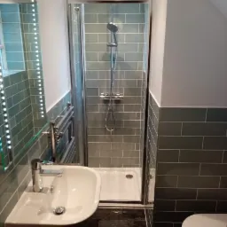 Nowoczesna łazienka z kabiną prysznicową wyłożoną szarymi płytkami, umywalką na drewnianym blacie, grzejnikiem drabinkowym i podświetlanym lustrem.