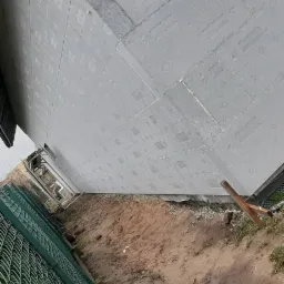 Ściana budynku pokryta szarym styropianem z widocznymi punktami kleju, przygotowana do dalszych prac wykończeniowych. Ziemia wokół budynku przygotowana, widoczna siatka ogrodzeniowa i fragment...