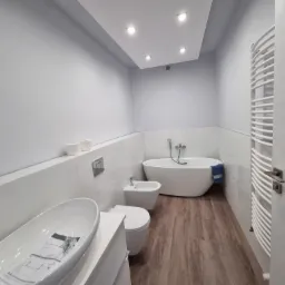 Nowoczesna łazienka w odcieniach szarości i bieli z wolnostojącą wanną, podwieszaną toaletą i bidetem, umywalką nablatową oraz grzejnikiem drabinkowym, oświetlona punktowymi lampami w suficie.
