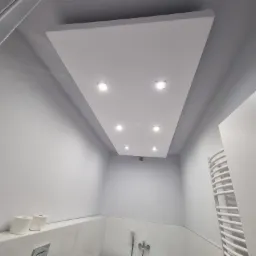 Nowoczesna łazienka w odcieniach szarości i bieli z podwieszanym sufitem z oświetleniem punktowym LED, widoczny grzejnik drabinkowy, armatura i akcesoria łazienkowe.