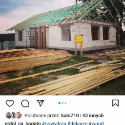 Surowy stan domu z białego bloczka z częściowo wykonaną konstrukcją dachu z zielonych belek, deski leżące na ziemi, tablica informacyjna na elewacji, widok na las w tle.