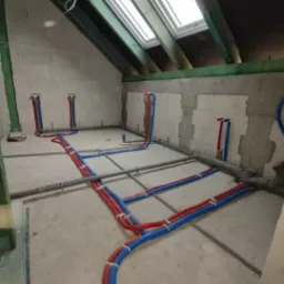 Poddasze w trakcie remontu z widoczną instalacją hydrauliczną: czerwono-niebieskie rury poprowadzone po podłodze i w ścianach, szare rury kanalizacyjne, okno dachowe w skośnym suficie.