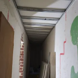 Wąski korytarz w trakcie prac wykończeniowych, widoczna konstrukcja metalowa podwieszanego sufitu z płyt kartonowo-gipsowych, ściany z nierównościami i śladami farby, w tle sterta płyt i wiaderka...