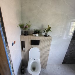 Wawrzynowicz - Nowoczesna łazienka z podwieszaną toaletą, zabudową z pionowych lameli i roślinami, wykończona szarymi płytkami marmurowymi. Widoczny fragment ościeżnicy z pianką montażową po lewej stronie...