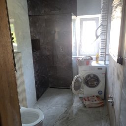 Wawrzynowicz - Nowoczesna łazienka z kabiną walk-in wyłożoną ciemnymi płytkami imitującymi marmur, jasnymi płytkami na podłodze i ścianach, pralką i podwieszaną toaletą. Sufit w surowym stanie.