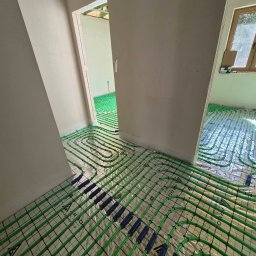 Wawrzynowicz - Zielone rury ogrzewania podłogowego ułożone na odblaskowej izolacji w kilku pomieszczeniach, z widocznymi przejściami i nieukończonymi ścianami.