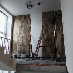 Wawrzynowicz - Wnętrze w trakcie wykańczania: ściany z drewnopodobnymi płytkami ułożonymi w jodełkę, z widocznymi klinami dystansowymi. Na pierwszym planie szare schody i drewniana drabina.