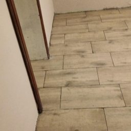 Wawrzynowicz - Podłoga wyłożona beżowymi płytkami imitującymi drewniane deski, ułożonymi z przesunięciem, w narożniku jasnego pomieszczenia z widoczną ciemną ościeżnicą drzwiową i niedokończonymi ścianami.