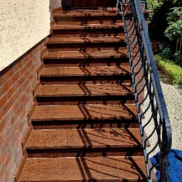 Zewnętrzne schody wejściowe wyłożone brązowym granitem, z czarną, ozdobną balustradą, prowadzące do drewnianych drzwi wejściowych z numerem 21 nad nimi.