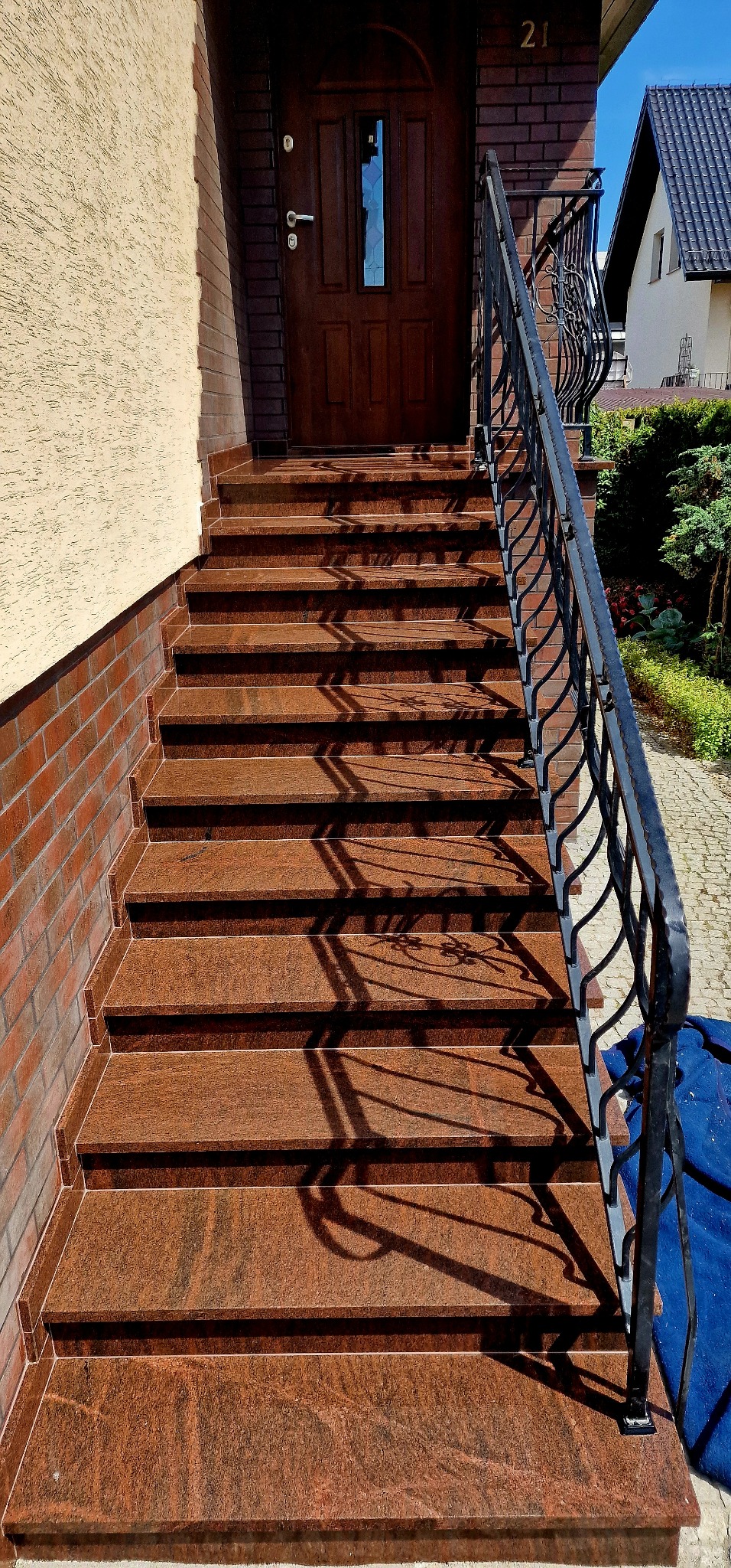 Zewnętrzne schody wejściowe wyłożone brązowym granitem, z czarną, ozdobną balustradą, prowadzące do drewnianych drzwi wejściowych z numerem 21 nad nimi.