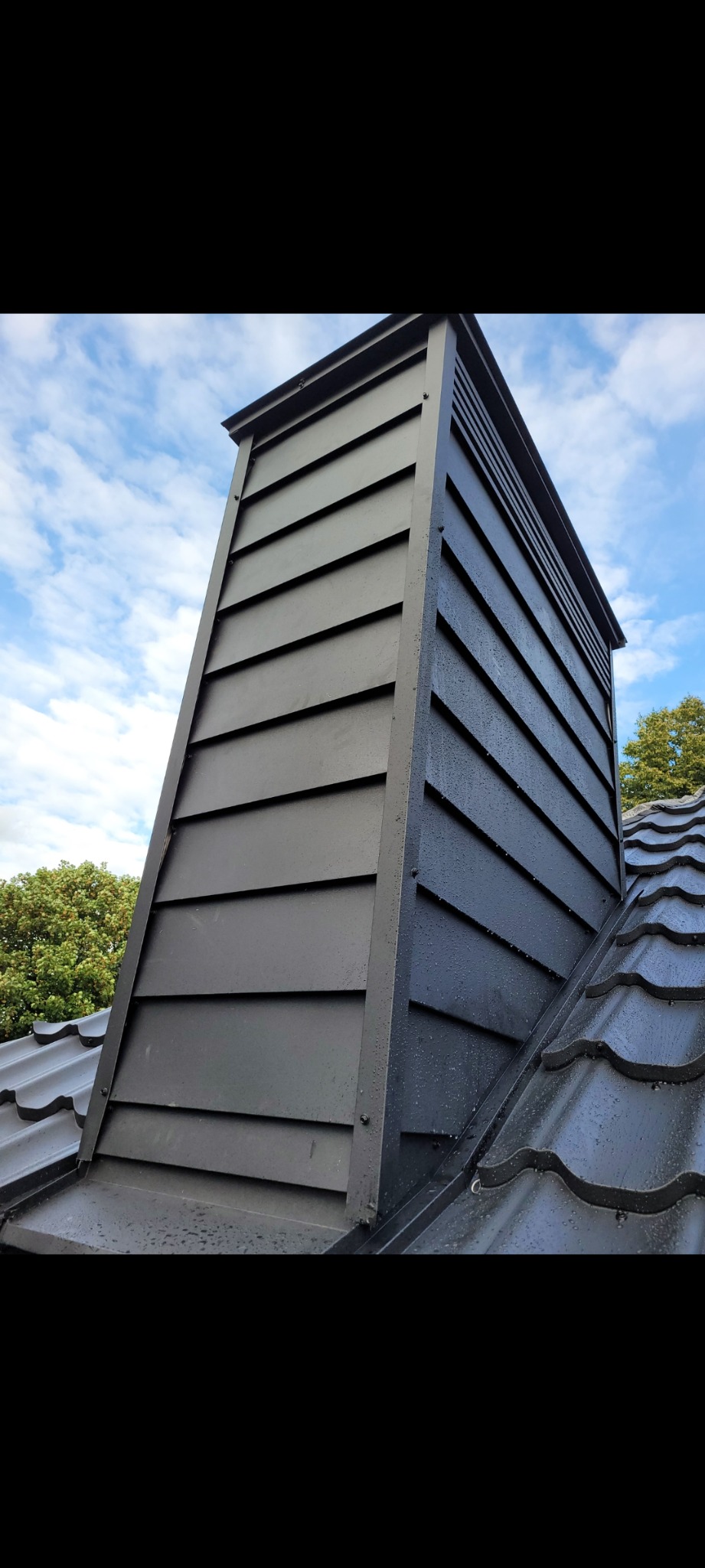 Nowoczesny, grafitowy komin wentylacyjny z poziomymi panelami, zamontowany na ciemnym dachu z dachówki ceramicznej, widoczne krople deszczu, w tle błękitne niebo z chmurami i fragment drzewa.