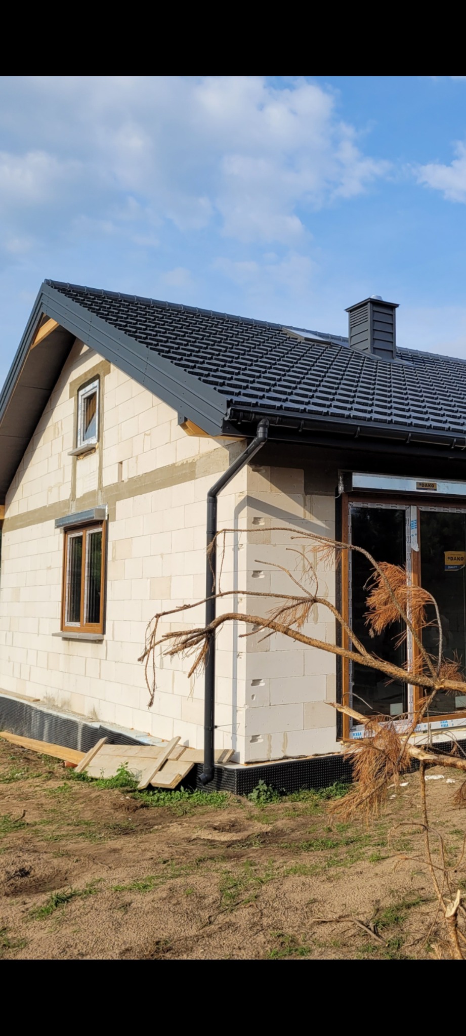 Nowy dom w trakcie budowy z szarą dachówką, widoczne okna z brązowymi ramami i rynna spustowa, na pierwszym planie uschnięta gałąź