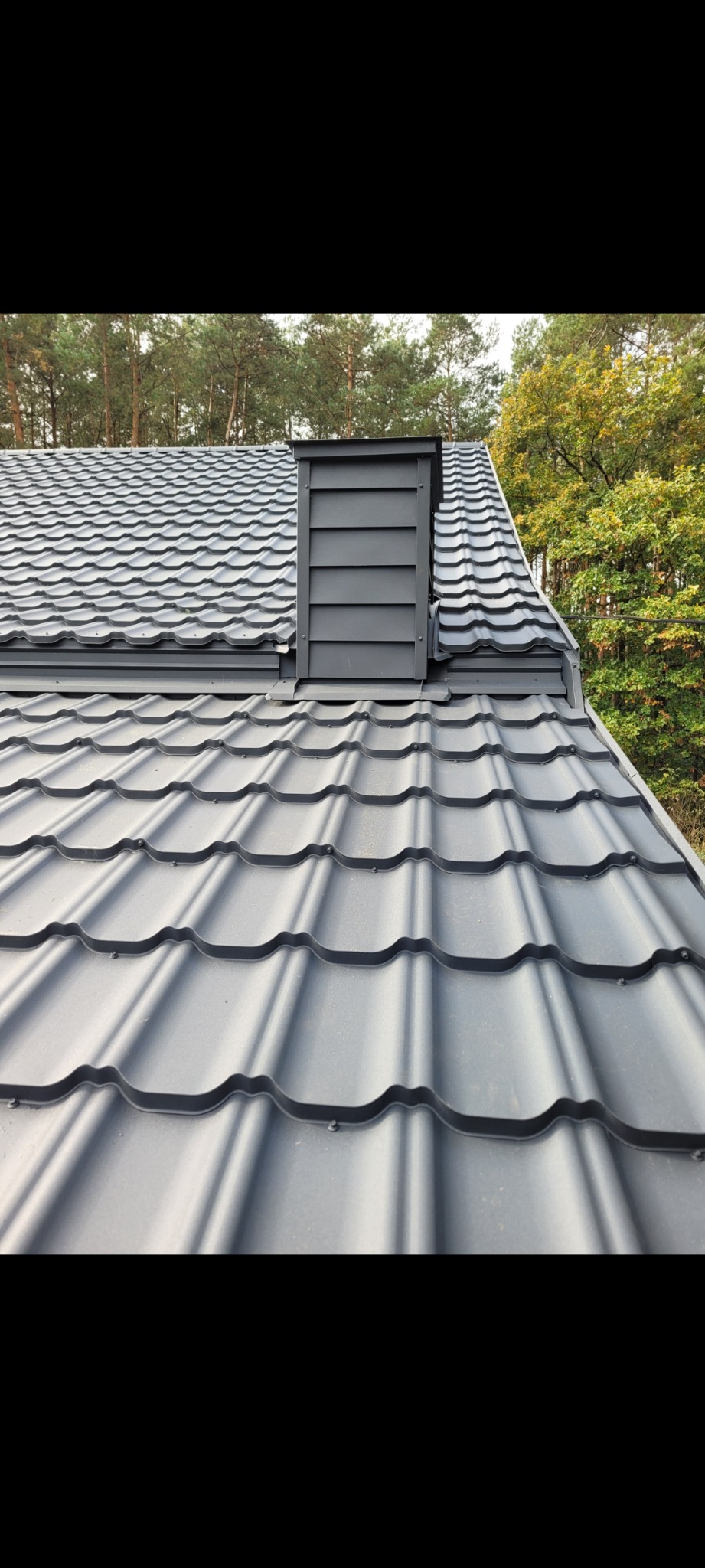 Szary dach z blachy trapezowej z kominem wentylacyjnym na tle drzew