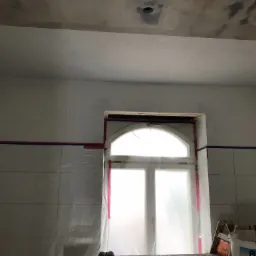 Przygotowania do prac wykończeniowych w łazience: zabezpieczone okno folią, taśmy malarskie na kafelkach, widoczne ślady gładzi na suficie i ścianach, wiadro z farbą marki Brillux.