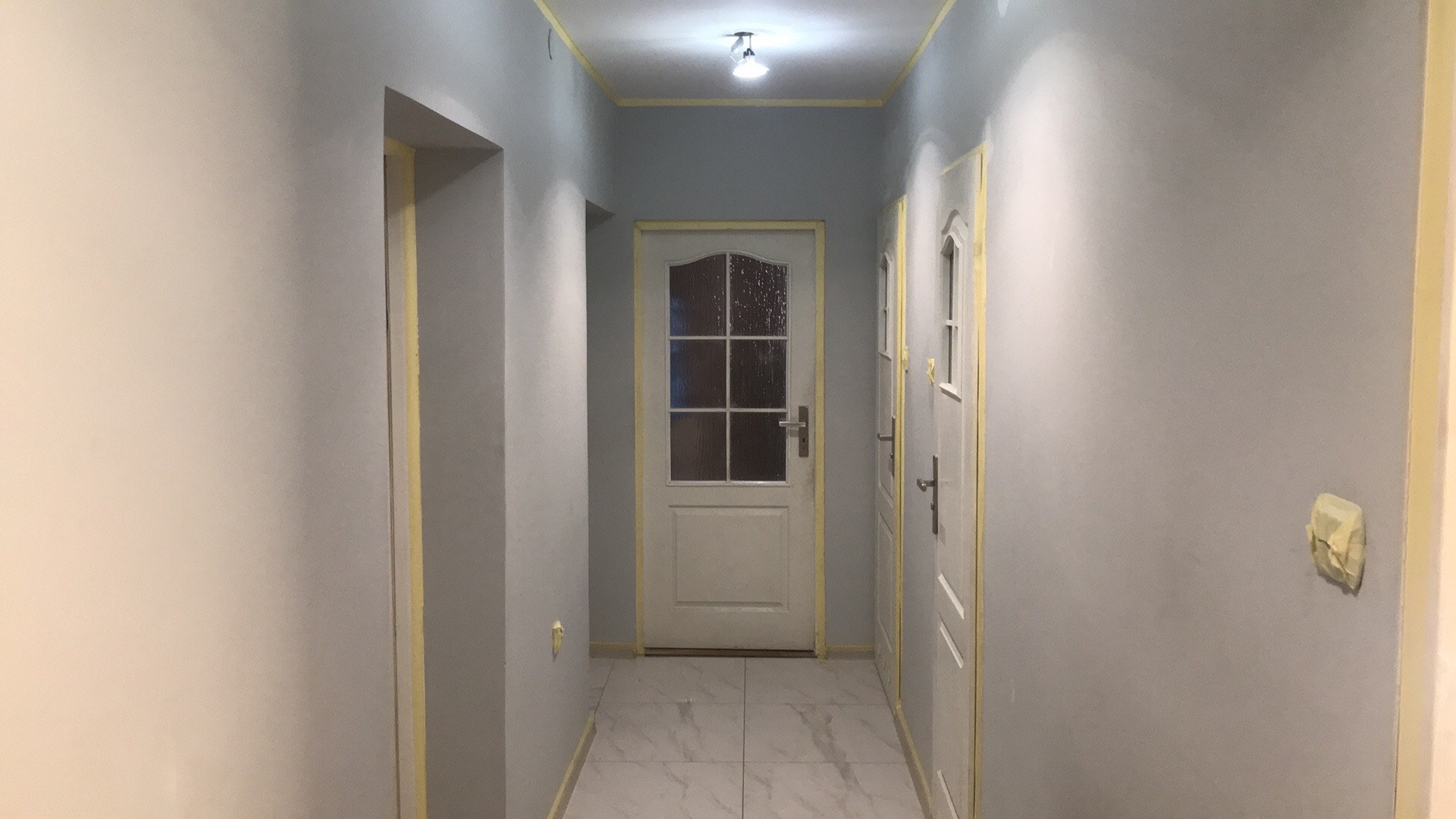 Korytarz w trakcie malowania, ściany w odcieniach szarości, zabezpieczone taśmą malarską, widoczne drzwi z szybami i marmurowe płytki na podłodze.