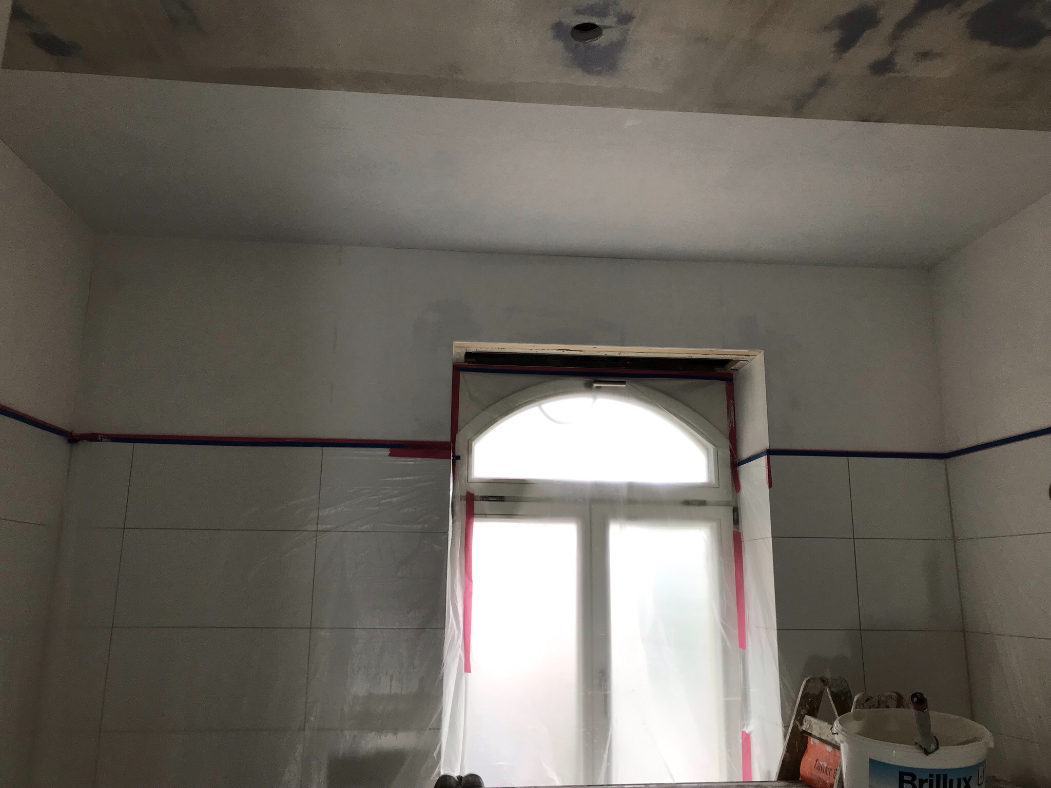Przygotowania do prac wykończeniowych w łazience: zabezpieczone okno folią, taśmy malarskie na kafelkach, widoczne ślady gładzi na suficie i ścianach, wiadro z farbą marki Brillux.