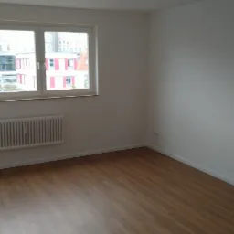 Pusty pokój po remoncie z panelami podłogowymi, białymi ścianami i grzejnikiem pod oknem z widokiem na budynek biurowy.