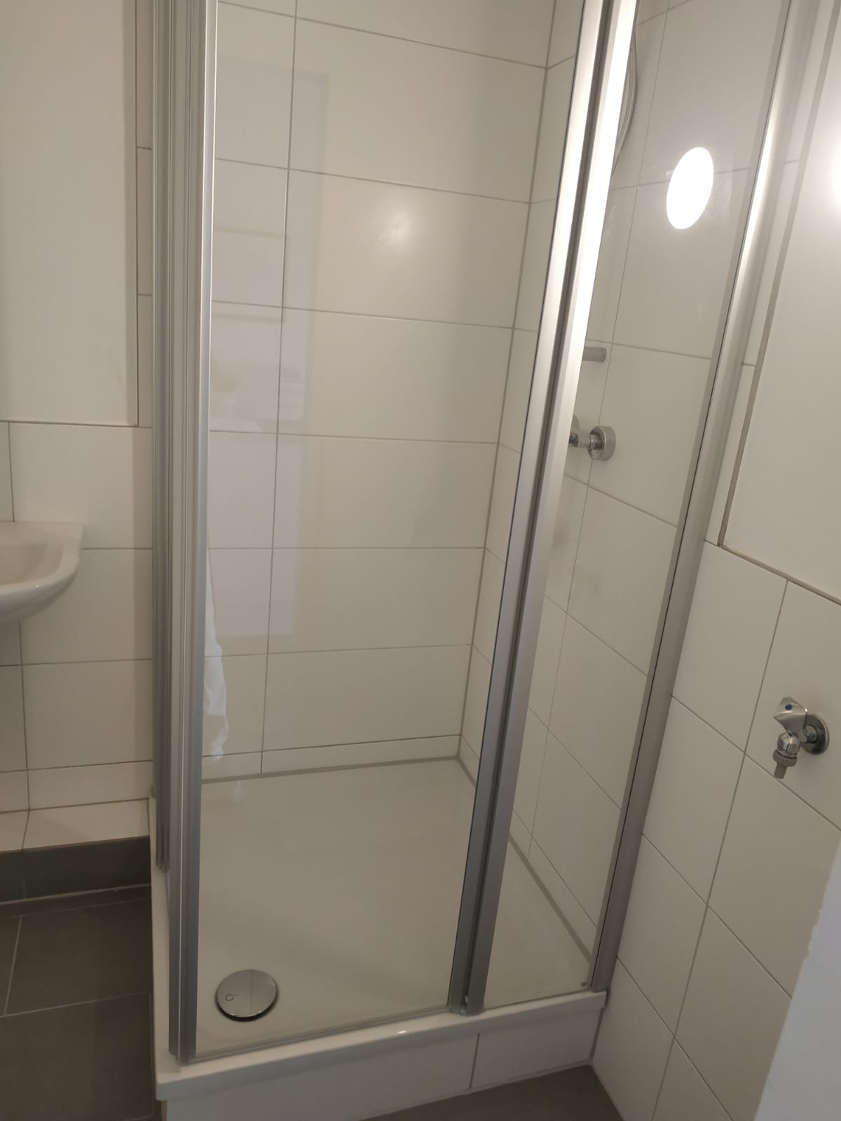 Nowoczesna kabina prysznicowa z chromowanymi profilami i białymi płytkami, widoczny odpływ liniowy i bateria umywalkowa w tle.