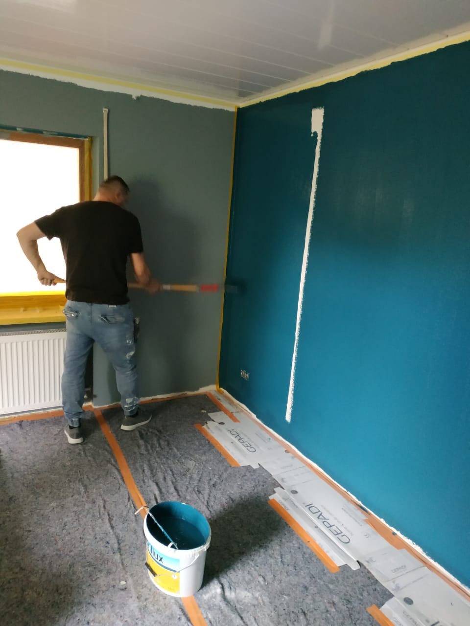 Mężczyzna maluje ścianę na niebiesko wałkiem, zabezpieczona podłoga, widoczny kubeł z farbą Dulux i taśma malarska.