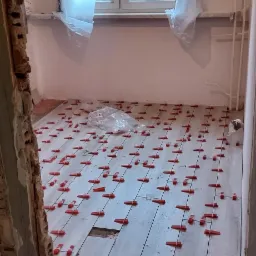 Podłoga w trakcie kładzenia płytek imitujących drewno, z użyciem czerwonych klipsów poziomujących, widoczny fragment ściany z odsłoniętą cegłą, okno zabezpieczone folią malarską.