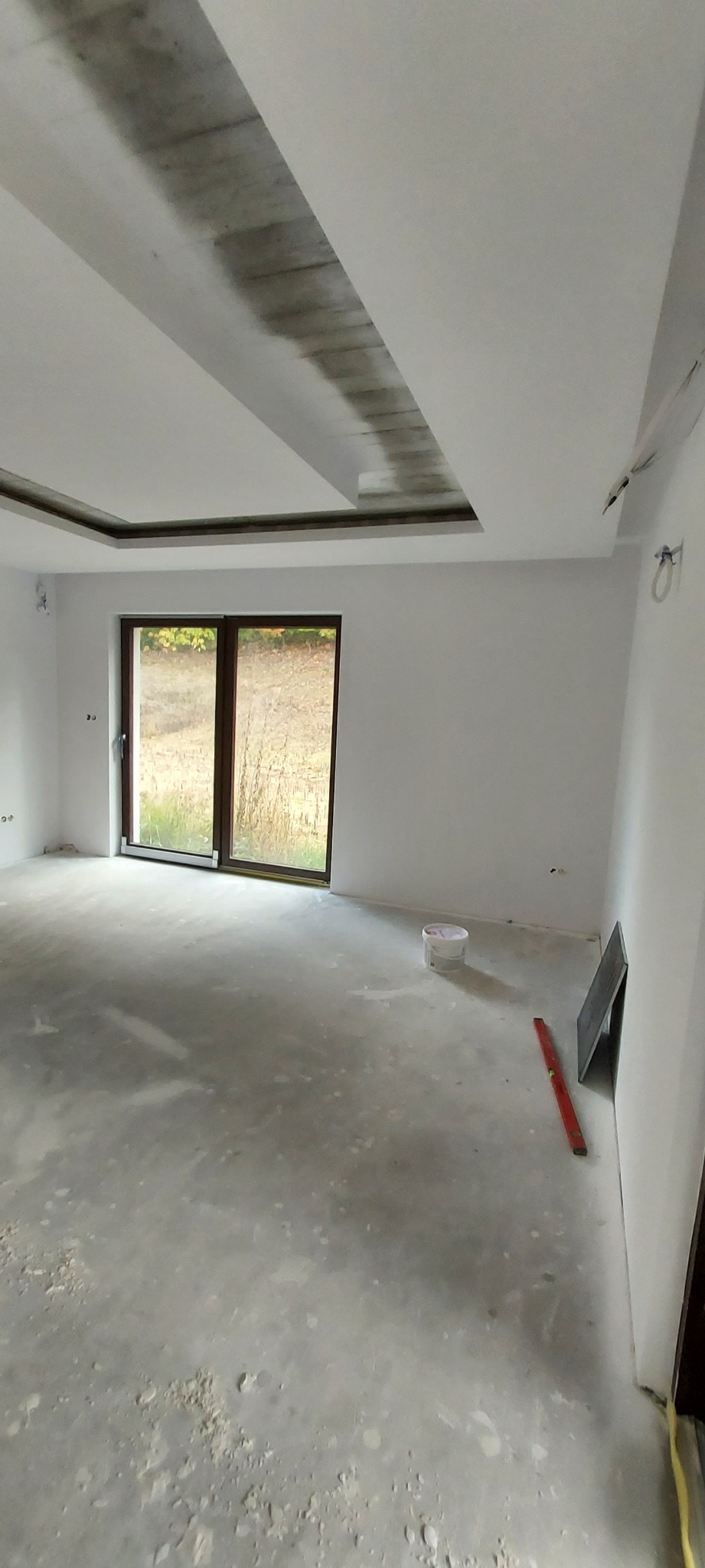 Wnętrze w trakcie prac wykończeniowych: surowy sufit z widoczną konstrukcją, białe ściany, betonowa podłoga, okno balkonowe, poziomica i wiadro z farbą.