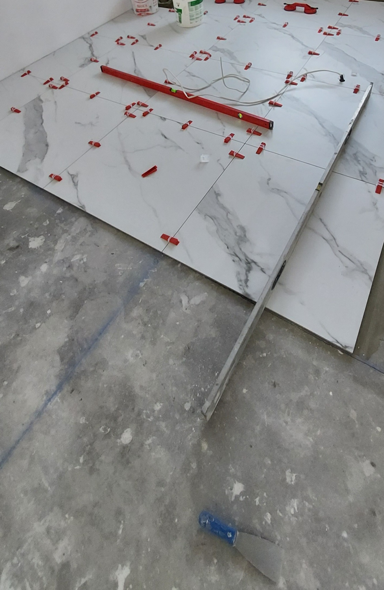 Fragment podłogi w trakcie układania płytek imitujących marmur, z użyciem poziomicy, krzyżyków dystansowych i szpachelki z niebieską rączką, widoczne ślady po kleju na podłożu.