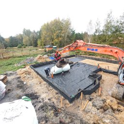 P.W. AS-KOP USŁUGI BUDOWLANO TRANSPORTOWE Albert Słomiński - Fundamenty domu w trakcie budowy: wykop zabezpieczony czarną folią, pracownicy i koparka w akcji. Widok z góry na plac budowy w otoczeniu drzew.