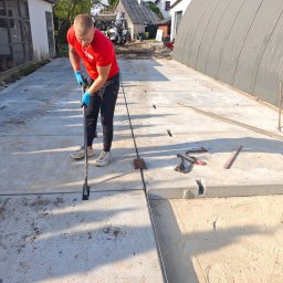 P.W. AS-KOP USŁUGI BUDOWLANO TRANSPORTOWE Albert Słomiński - Pracownik w czerwonej koszulce wyrównuje betonowe płyty z wycięciami, tworzące utwardzoną powierzchnię. W tle budynki i sprzęt budowlany.