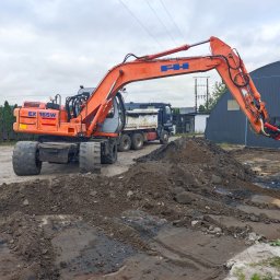 P.W. AS-KOP USŁUGI BUDOWLANO TRANSPORTOWE Albert Słomiński - Pomarańczowa koparka kołowa FH EX 165W w trakcie prac ziemnych, z widoczną stertą ziemi i ciężarówką w tle, na tle szarego nieba i zieleni.