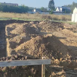 Przygotowanie terenu pod budowę: wykopane rowy pod fundamenty, pryzmy ziemi, drewniane szalunki i bloczki betonowe w tle.