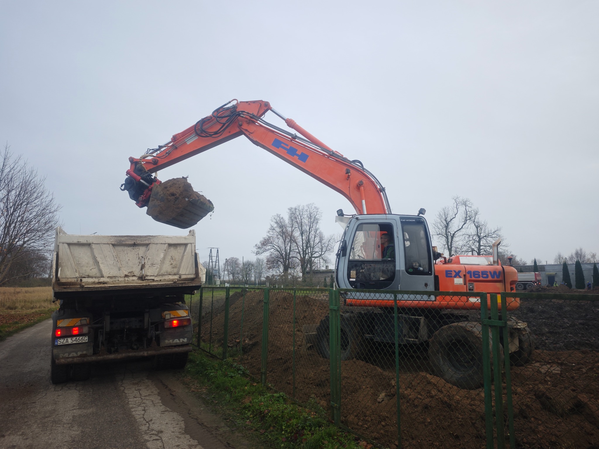 Pomarańczowa koparka nabiera ziemię do wywrotki MAN na tle zielonego ogrodzenia i pochmurnego nieba. Prace ziemne w toku, widoczny operator w kabinie.